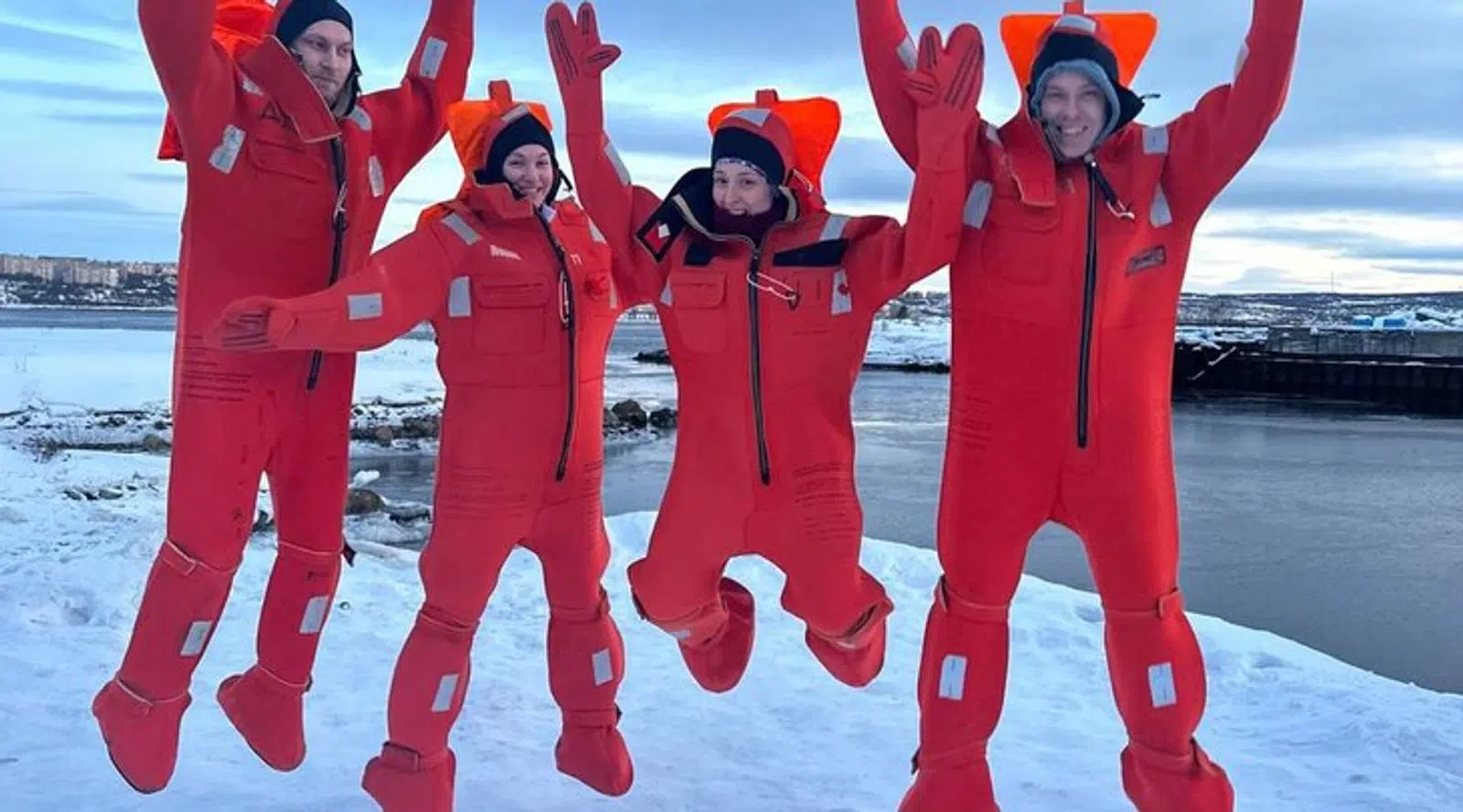 Four travelers jumping in immersion suits on the snowy shore